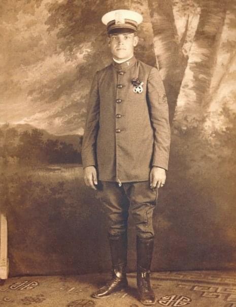 Sepia studio portrait of Chuck Credo Sr. as a young man, standing in a buttoned uniform jacket, peaked cap, breeches and tall boots before a painted landscape backdrop.