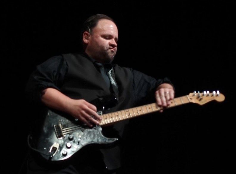 Chuck Credo IV playing a black Stratocaster-style guitar on a dark stage, head tilted down into the solo.