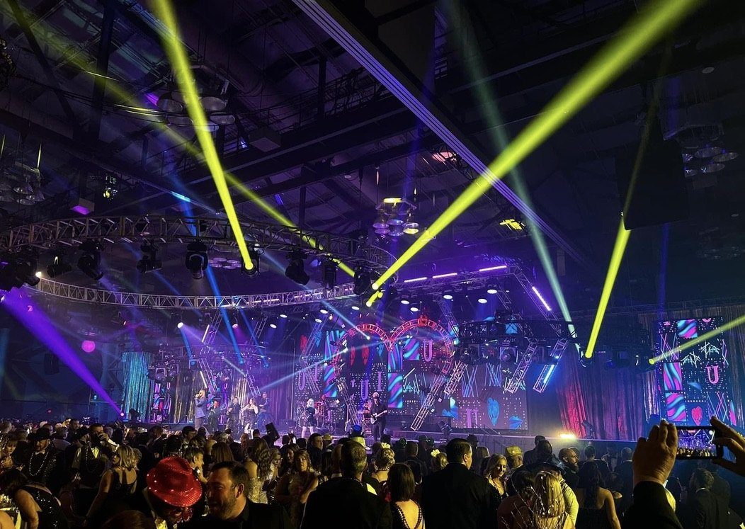 A convention hall stage with yellow and purple moving-light beams over a large crowd.