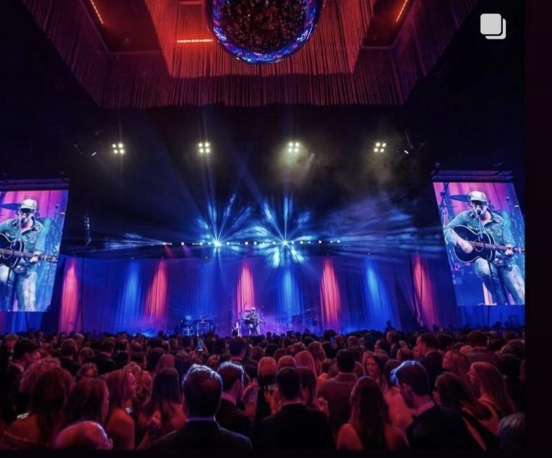 A large ballroom audience facing a lit stage, with live video of the band on screens either side.