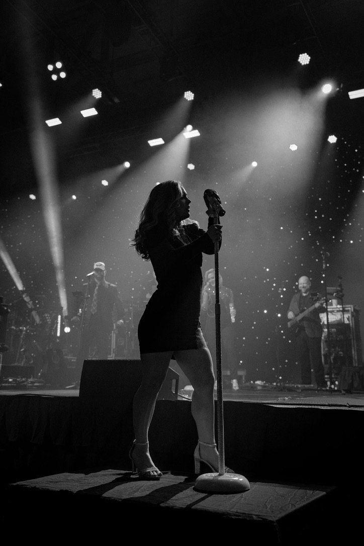Black-and-white photo of a vocalist singing at the lip of a large stage, silhouetted by spotlights.