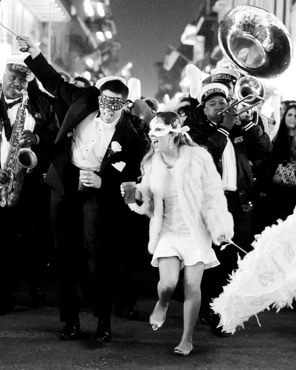 Black-and-white photo of a second line in full swing: a masked couple dancing, a sousaphone and a feathered fan behind them.
