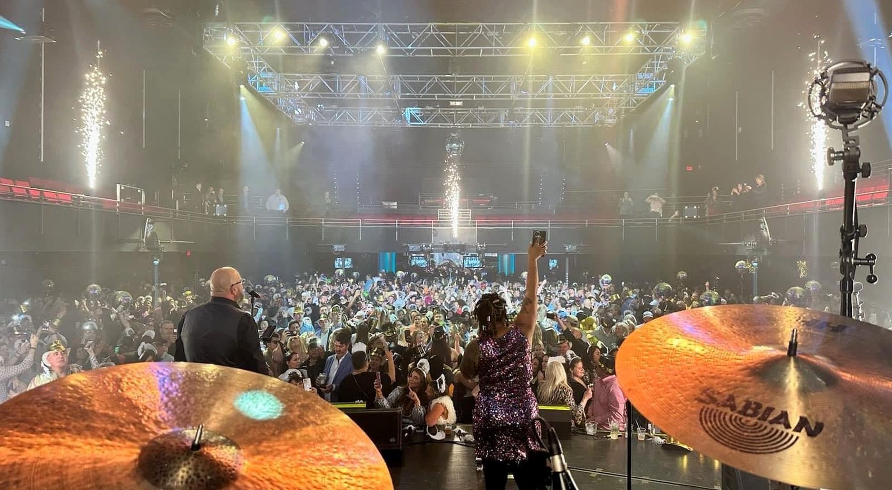 The view from behind the drum kit at a big show: cymbals in the foreground, a singer at the front of the stage, and a huge crowd under lighting truss.