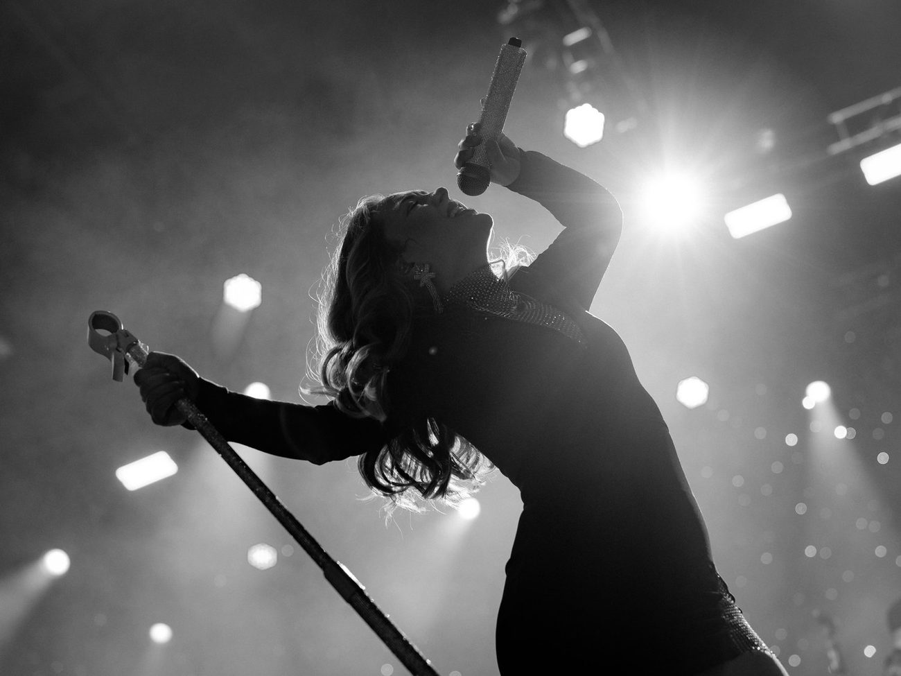 Black-and-white photo of a vocalist leaning back with her microphone held high, backlit by stage lights.