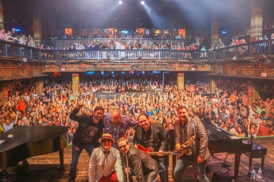 Billy vs Elton on stage after a show, the band posing between two grand pianos in front of a packed theatre, the floor and balcony full of raised hands.