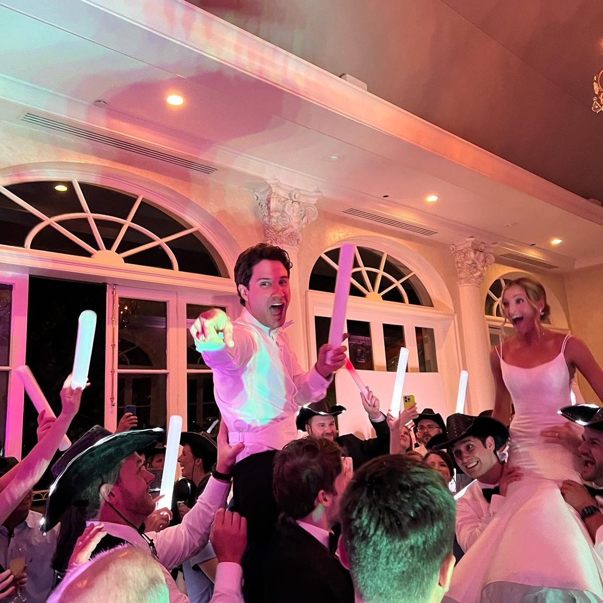 A bride and groom lifted up by their guests, waving light-up batons under pink lighting.