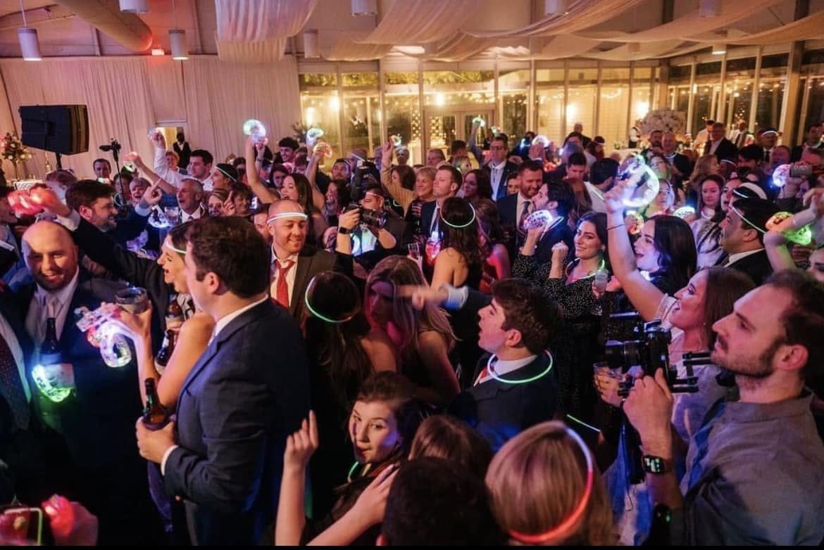 A packed wedding dance floor, guests in suits and dresses waving glow sticks.
