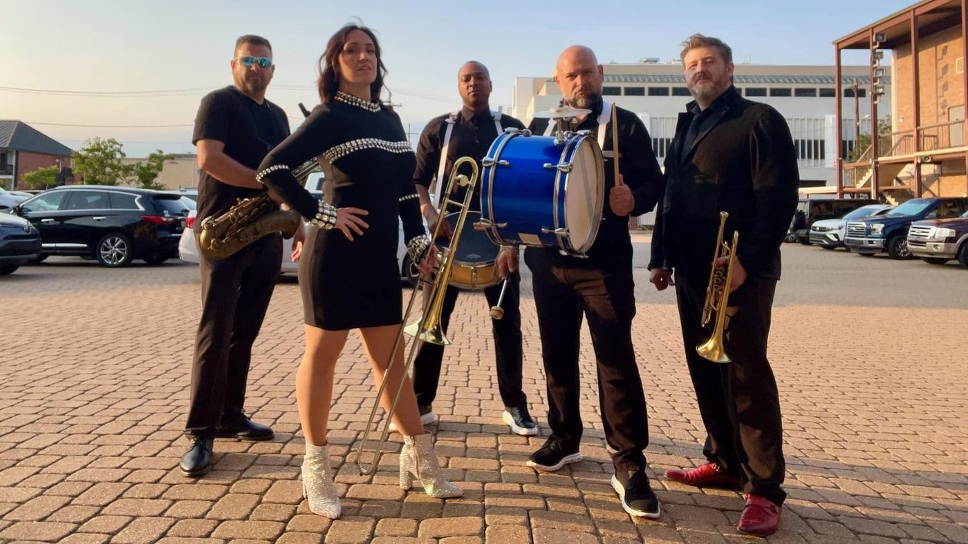 A brass band — trumpet, trombone, saxophone and bass drum — lined up outside at golden hour, ready for a second line.