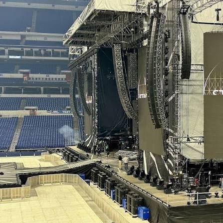 A stadium stage build seen from the seating deck — flown line array speaker hangs, lighting truss, LED screens and stage decking in an empty arena before doors.